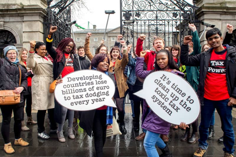 Protesters holding signs against tax dodging, advocating for fair tax systems, outside a historic building.