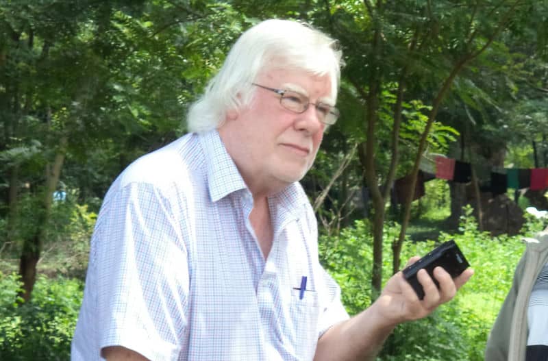 Man with white hair in a plaid shirt holding a device, standing outdoors surrounded by greenery.