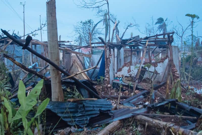 Severe hurricane damage to a house, with debris and fallen trees scattered around the collapsed structure.