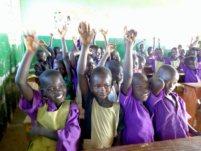Children in a classroom raising hands, wearing purple uniforms, smiling, and engaging in an educational setting.