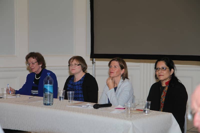 Four women speakers at a conference panel sitting at a table with microphones and water bottles.
