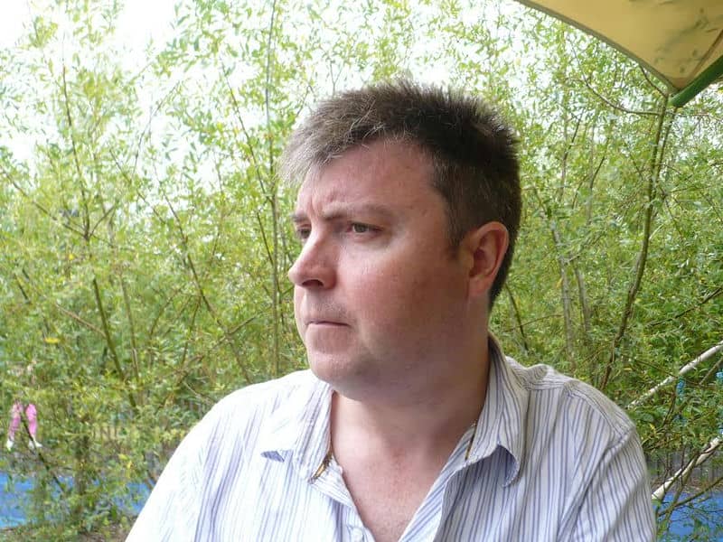 Man in striped shirt sitting outdoors near green foliage, gazing thoughtfully to the side.