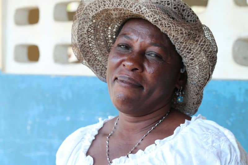 Woman in a straw hat and white blouse smiling, with a textured blue wall in the background.