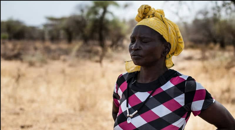Woman in patterned dress and yellow headwrap standing in dry, rural landscape.