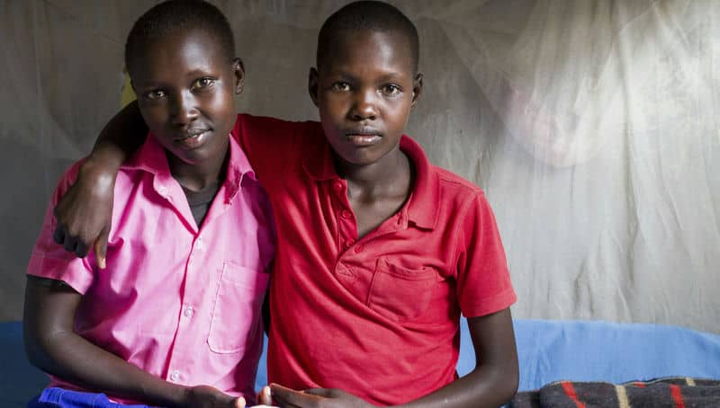 Two friends in colorful shirts sitting together, embracing in a simple room, conveying warmth and connection.