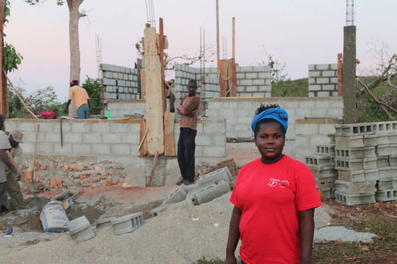 Woman in red shirt stands in front of a brick construction site with workers building walls in the background.