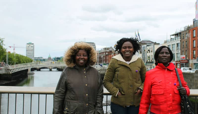 Three people in winter coats smiling on a bridge overlooking a river and cityscape.