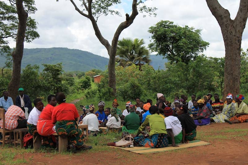 Outdoor community meeting in a rural setting, people gathered under trees with mountains in the background.