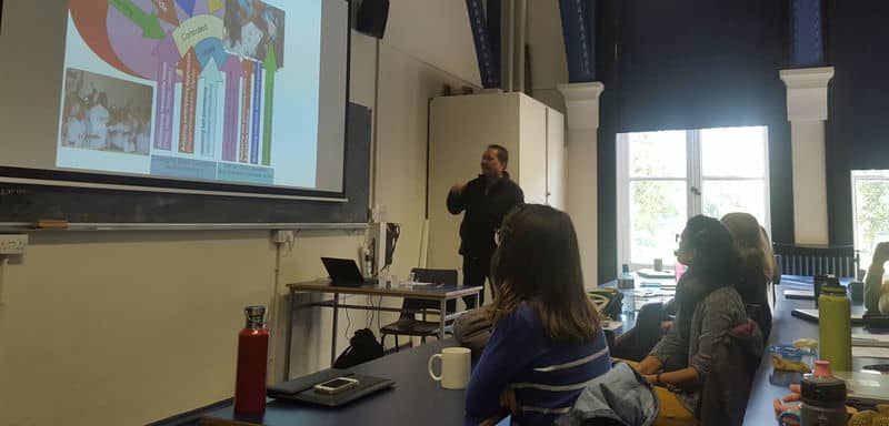 Classroom lecture with teacher presenting colorful chart to attentive students, natural light filters through large windows.