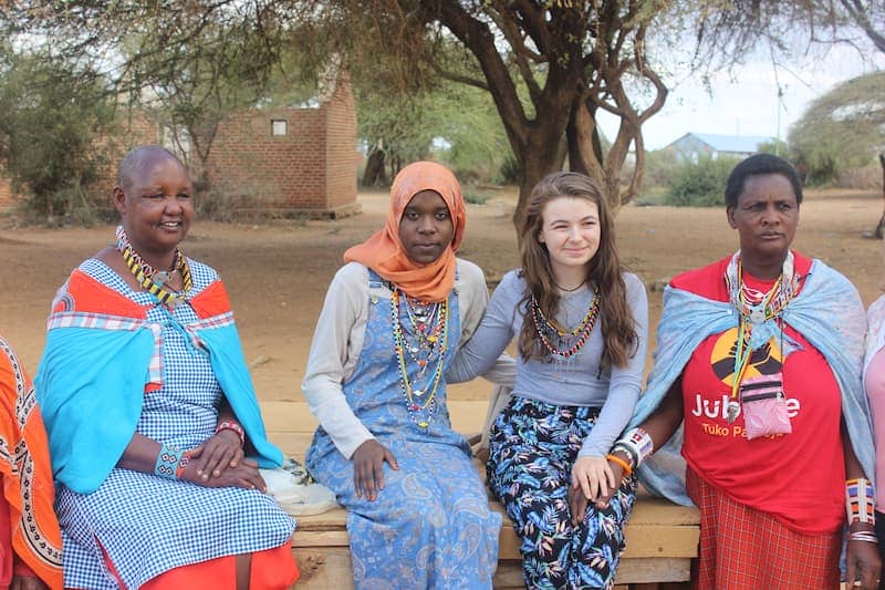 Group of women in colorful attire sitting on a bench outdoors, showcasing traditional necklaces and clothing.