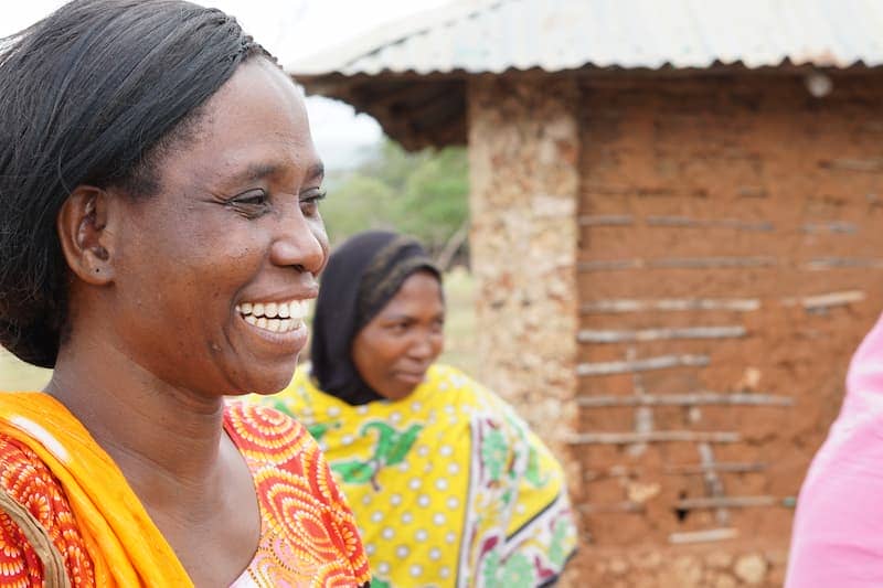 Woman smiling in vibrant clothing, standing outdoors near a traditional mud house.