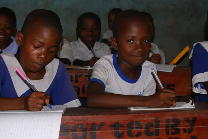 Children focused on writing in a classroom setting, wearing uniforms, and using notebooks and pens.