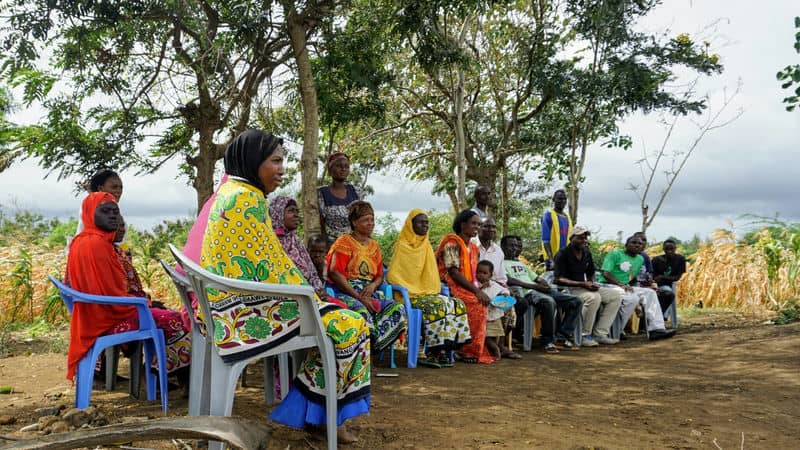 A community group sitting outdoors, engaged in discussion under trees with a rural landscape in the background.