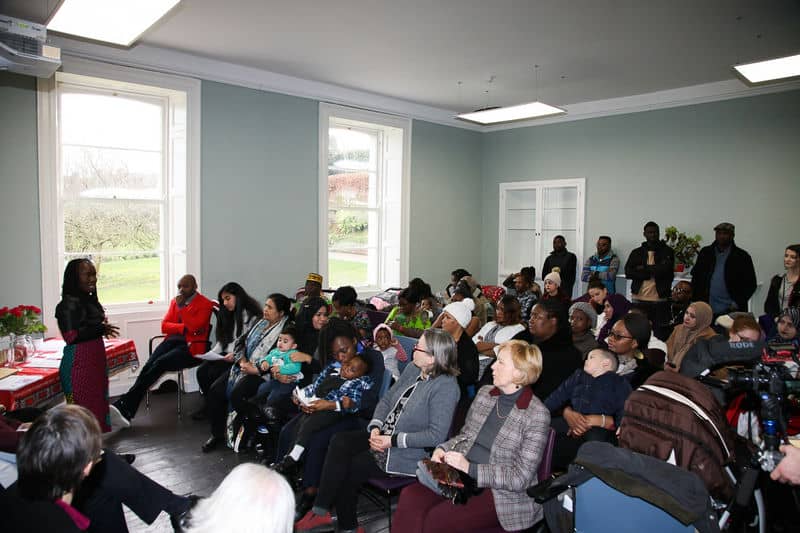 Community workshop with diverse group attentively listening to speaker in a bright room with large windows.