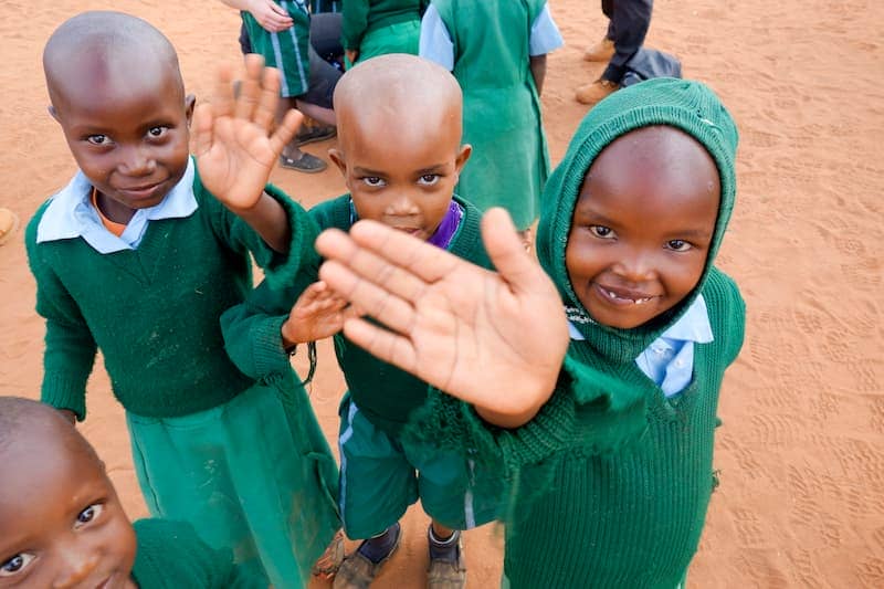 Children in green school uniforms smiling and waving at the camera in a sandy playground.