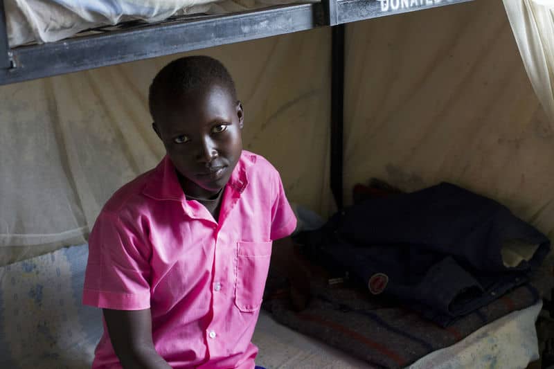 Child in a pink shirt sitting on a bunk bed in a simple room.