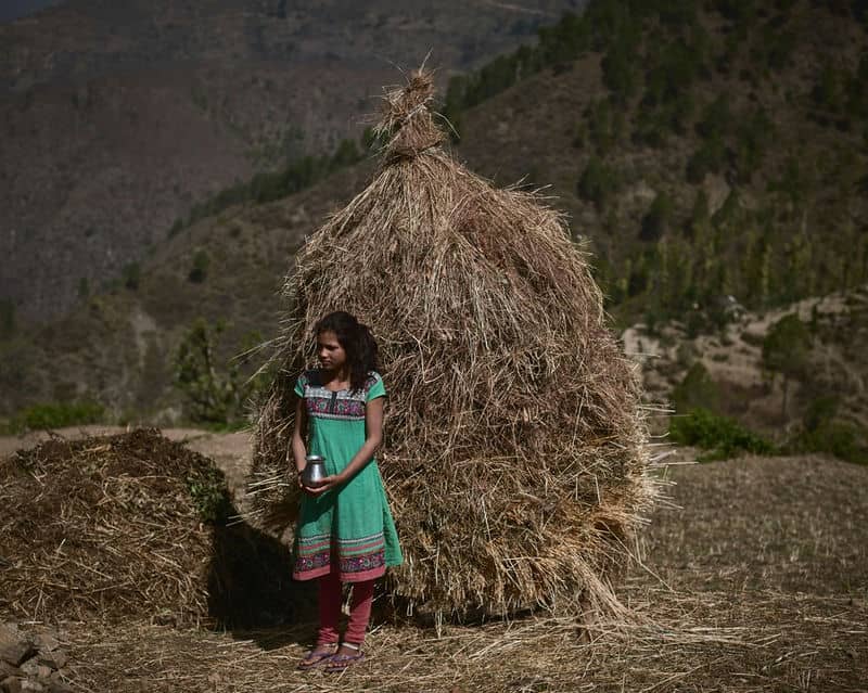 Young girl with a jug stands beside a haystack in mountainous rural landscape, wearing a colorful dress.