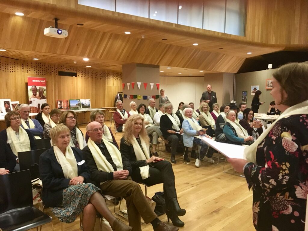 Audience at a formal gathering listens to a speaker in a wooden auditorium.