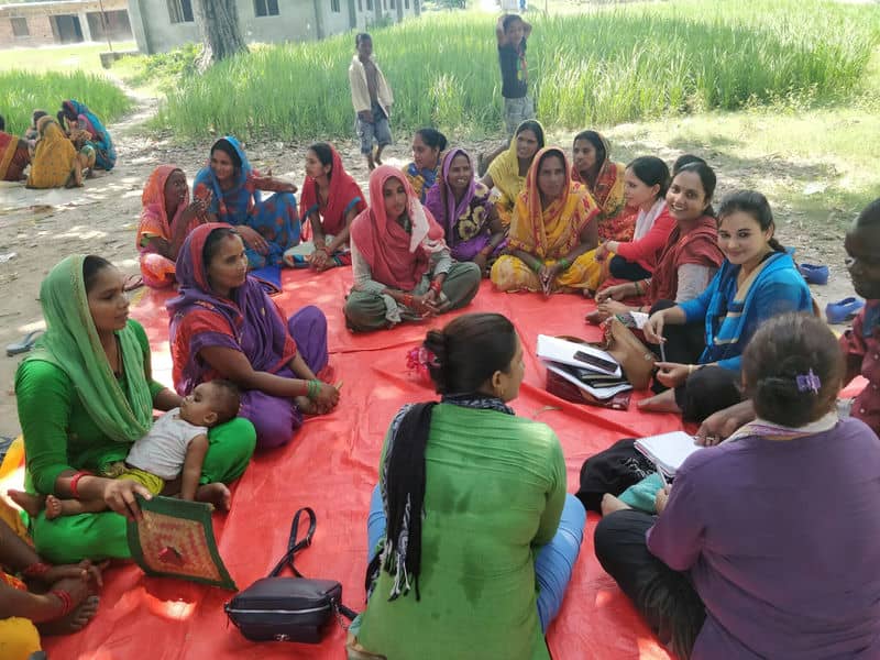 Group of women in colorful attire having a discussion outdoors on a red mat.