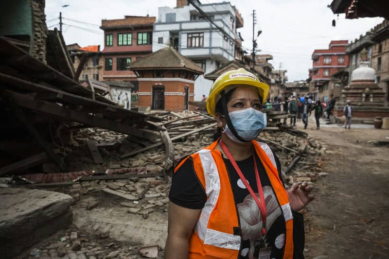 Construction worker in safety gear inspects rubble in an urban area wearing a mask, hard hat, and orange vest.