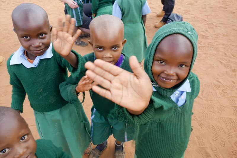 Smiling children in green school uniforms waving at the camera on a dirt playground.