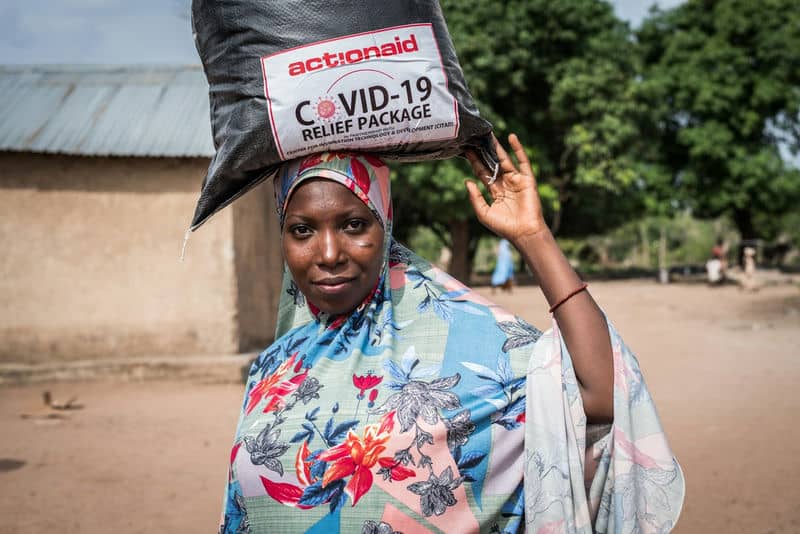 Woman carrying COVID-19 relief package on her head, wearing a colorful hijab, standing outside a rural house.
