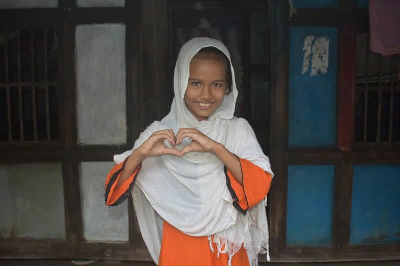 Smiling girl in white hijab and orange dress making a heart shape with hands in front of a rustic doorway.