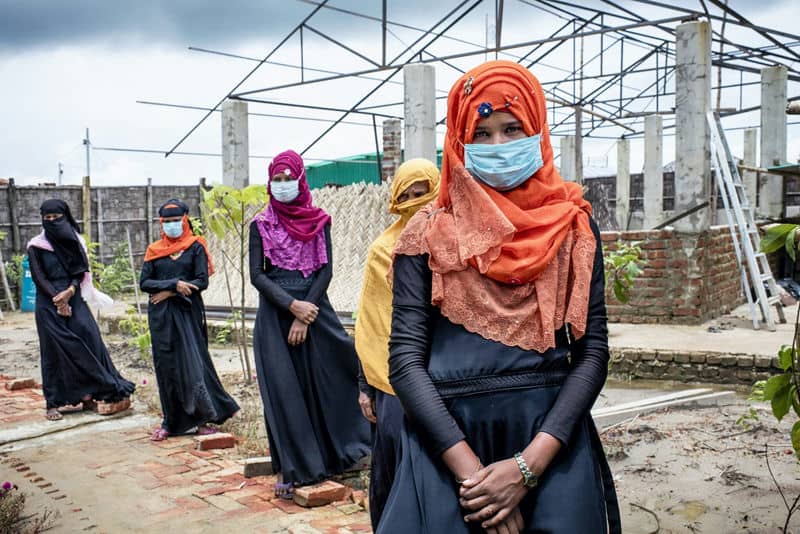 Group of women in colorful hijabs and masks standing in an outdoor construction area under a cloudy sky.