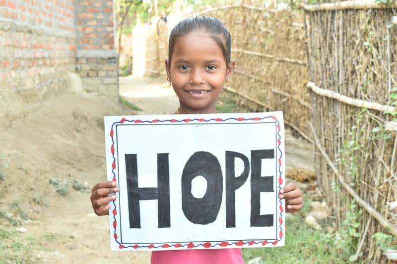 Girl holding a HOPE sign, smiling in a rural setting, symbolizing optimism and positivity.