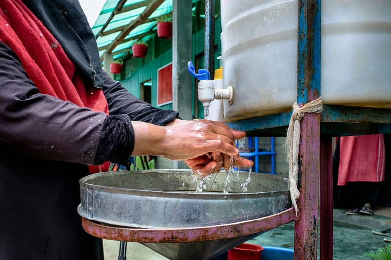 Person washing hands under a tap with a large water container, promoting hygiene and cleanliness outdoors.