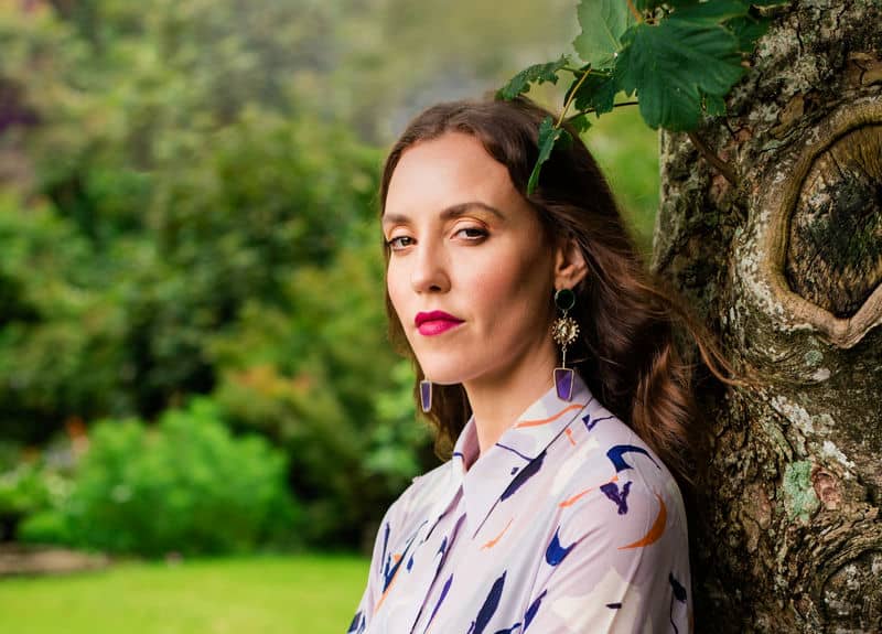 Woman with earrings poses against a tree in a garden wearing a patterned shirt.