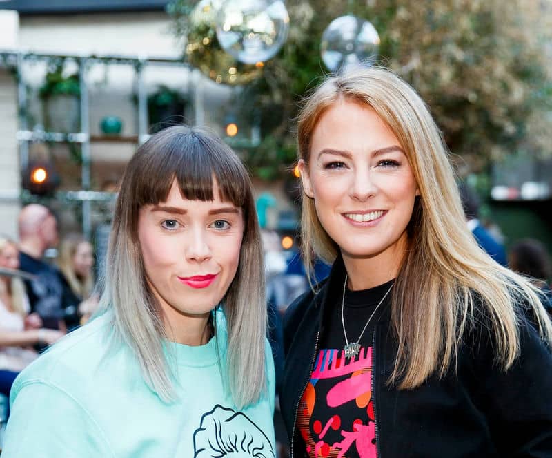 Two women smiling at an outdoor event with festive decorations in the background.