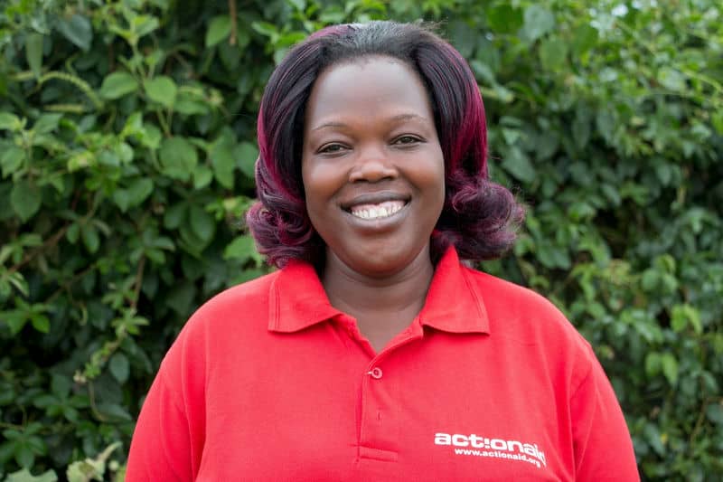 Portrait of a smiling woman in a red actionaid shirt, standing in front of green foliage.