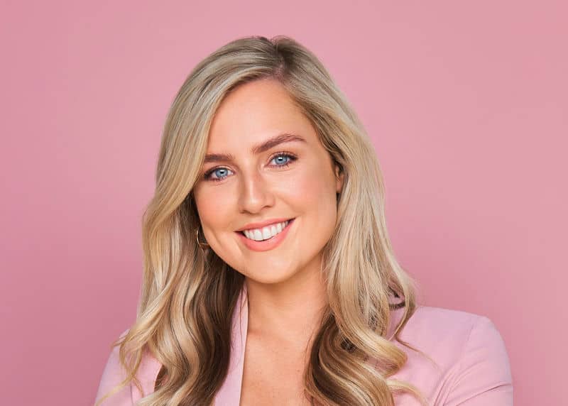 Smiling woman with long blonde hair in a pink blazer, set against a pink background.