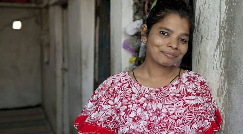 Smiling woman in a floral red dress leaning against a wall in a corridor.