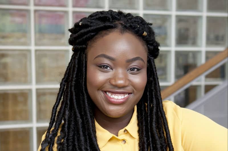 Smiling person with braided hair wearing a yellow shirt, standing in front of a glass-tiled wall.