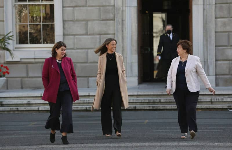 Three women walking outdoors, smiling, in front of a stone building with a man in the background.