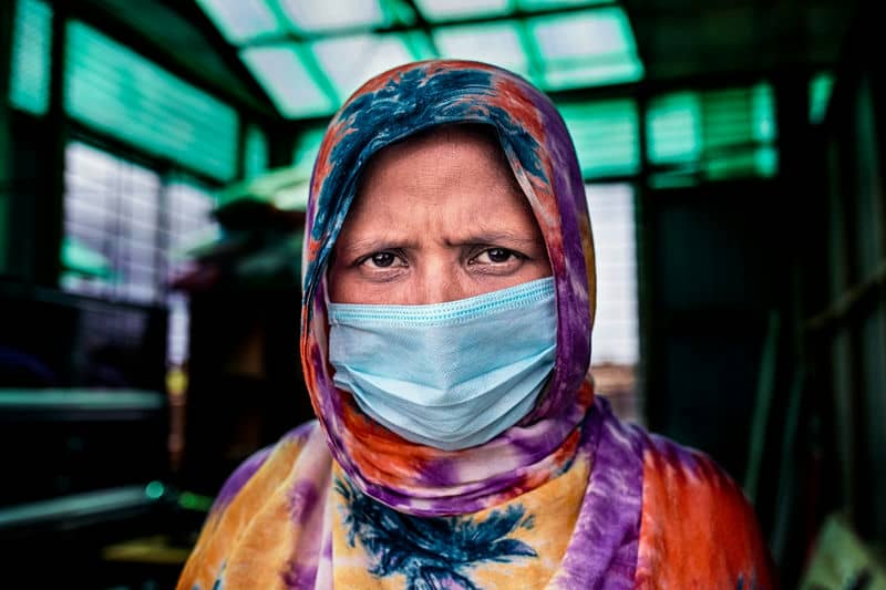 Woman in colorful hijab wearing a face mask, standing indoors, looking determined, against a blurred background.