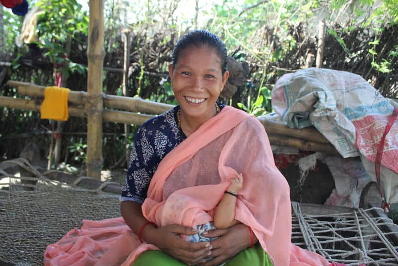 Smiling woman in pink saree breastfeeding a baby outdoors, sitting on a woven cot surrounded by greenery.