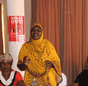 Woman in yellow hijab speaks at a community event, with Speak Out! poster in the background.