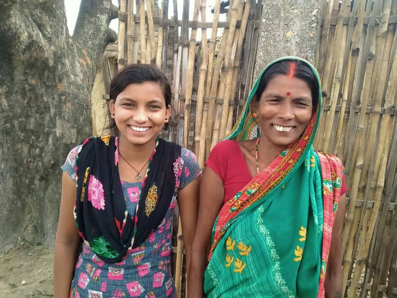 Two women standing and smiling in colorful traditional attire, against a bamboo and tree background.