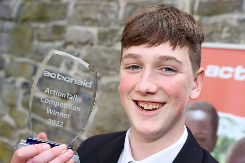 Person holding ActionAid ActionTalks Competition 2022 Winner trophy, smiling in front of a stone wall background.