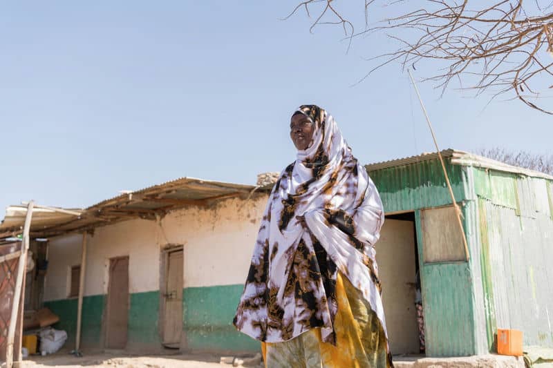 Woman in colorful attire stands outside rural house on a sunny day, showcasing traditional fashion and architecture.