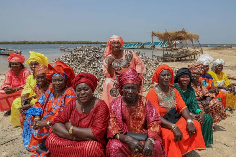 Group of women in vibrant traditional attire seated by riverbank with oyster shells, displaying community and culture.