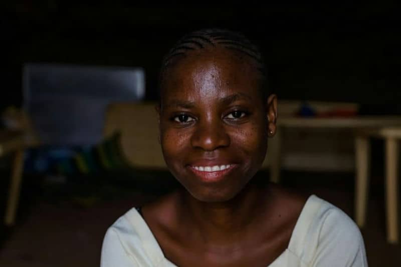 Smiling person with braided hair sitting indoors, featuring warm lighting and a friendly expression.