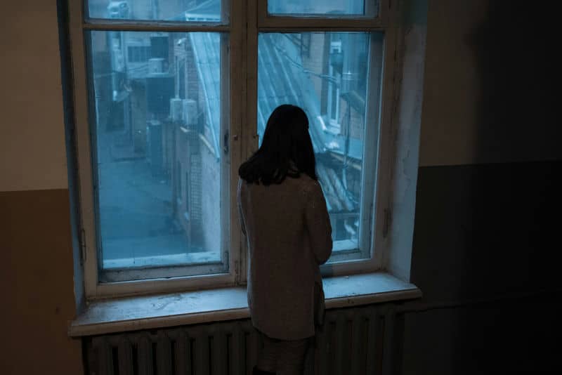 Person gazing through an old window at a cityscape, embraced by dim interior lighting.
