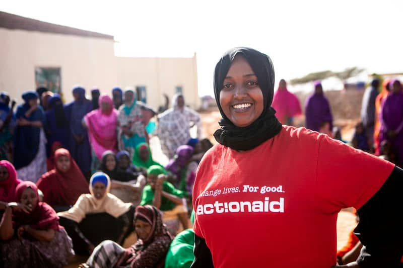 Smiling woman in ActionAid shirt with a community group in the background.