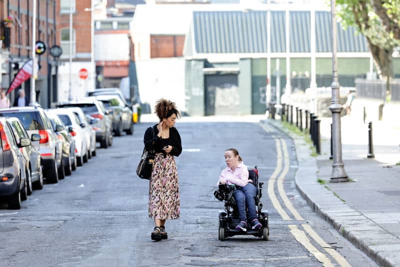 Two women, one using a wheelchair, chatting and walking down a city street. Urban accessibility theme.