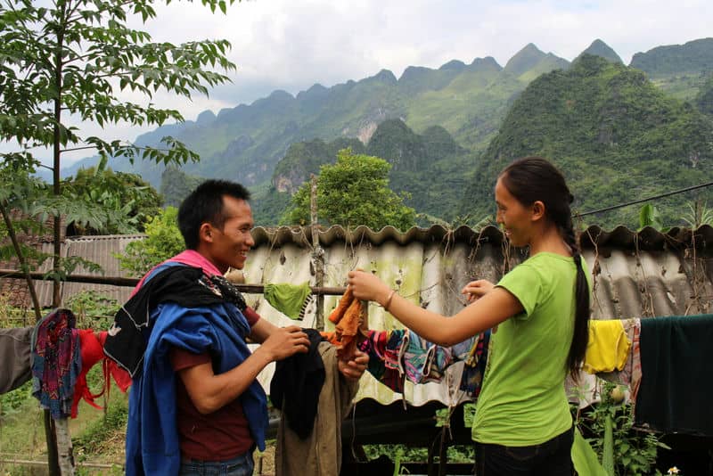 Two people hanging colorful clothes outdoors with lush green mountains in the background.