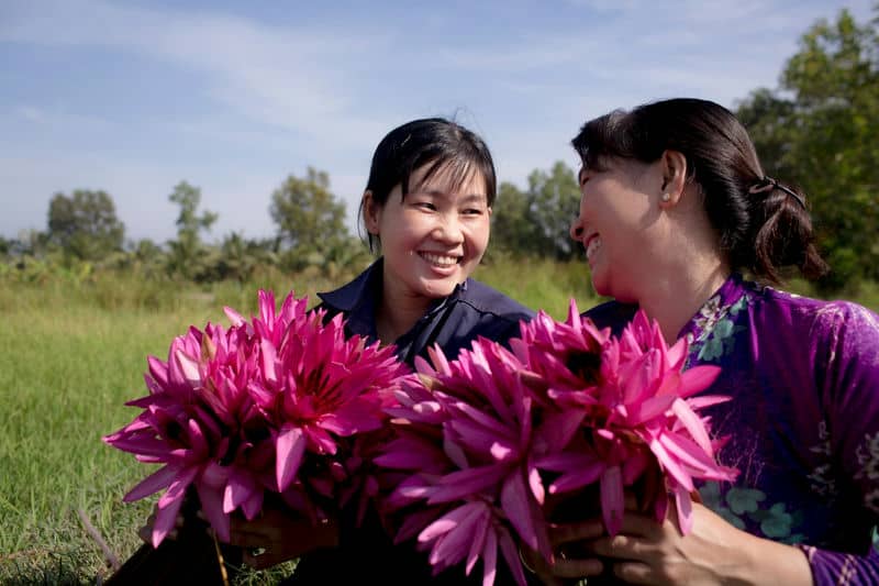 Two women joyfully holding vibrant pink water lilies in a sunny field, showcasing nature's beauty and friendship.